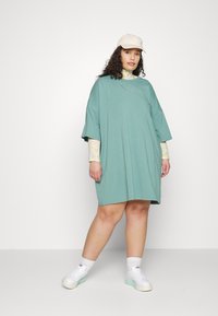 Vestido de algodão verde oversized com mangas curtas, combinado com uma camisa de manga longa estampada por baixo. A modelo usa uma cápsula bege e ténis brancos.