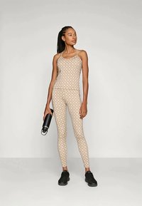 Γυναίκα με μπεζ καπιτονέ τοπ και κολάν κρατάει μαύρο μπουκάλι νερού, στέκεται με φόντο σε ανοιχτό φως, φορώντας μαύρα αθλητικά παπούτσια.