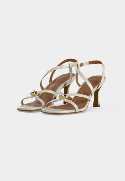 Paire de sandales blanches à talons hauts avec de fines lanières croisées et de petites décorations ovales dorées sur les lanières avant.