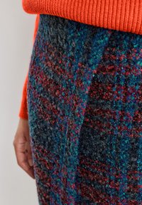 Zbliżenie osoby noszącej czerwony dziergany sweter oraz fakturowaną, wielokolorową wełnianą spódnicę z niebieskimi, czerwonymi i zielonymi wzorami.