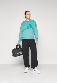 Teal hettegenser med en stor teal-logo, svarte vide bukser, hvite joggesko og en svart duffelbag med hvit merkevare. Uformell treningstøy.