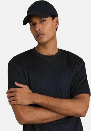 Marineblaues Baseballcap und schwarzes Shirt mit strukturiertem Wellenmuster. Das Shirt hat einen runden Halsausschnitt und kurze Ärmel und zeigt verschränkte Arme.
