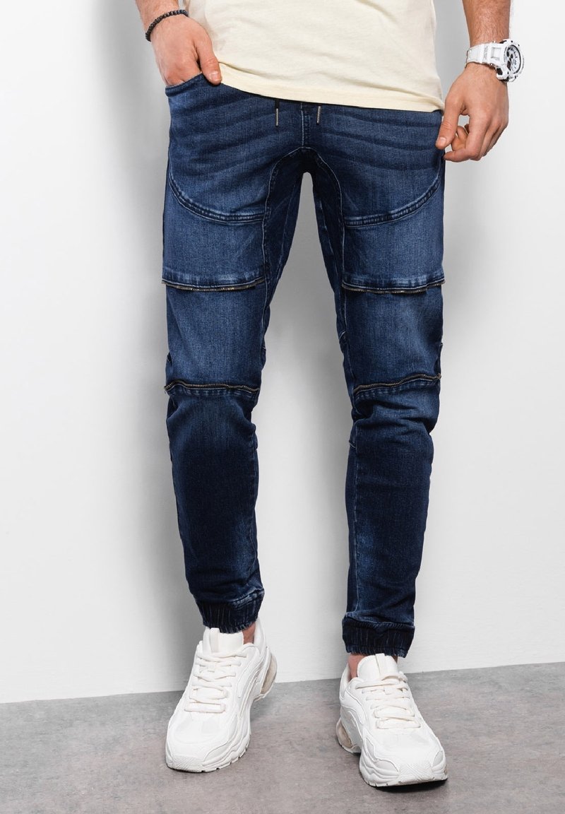 Homme portant un pantalon jogger en denim bleu foncé avec fermeture éclair et des baskets blanches, debout avec les mains près des poches contre un mur blanc.