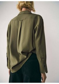 Chemise boutonnée texturée vert olive à coupe décontractée, manches longues, avec un détail d'empiècement au dos, portée avec un pantalon noir.