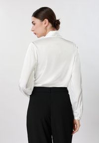 Femme aux cheveux foncés en chignon, vêtue d'un chemisier blanc à manches longues et d'un pantalon noir taille haute, tournée vers la gauche sur un fond uni.