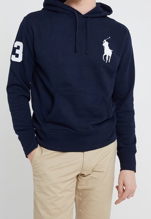 Mann trägt einen marineblauen Hoodie mit weißem Polo-Spieler-Logo auf der Brust und der Zahl 3 auf dem rechten Ärmel, kombiniert mit beigen Hosen.
