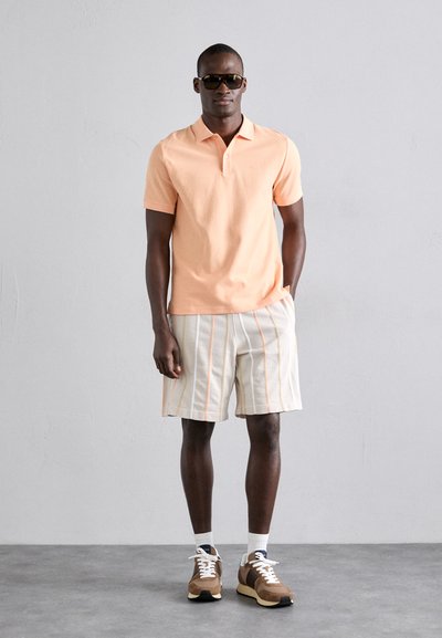 Lys orange polo shirt, beige shorts med lodrette striber i orange, brune sneakers med hvide snørebånd, solbriller.