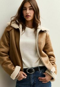 Giacca di shearling marrone con colletto marrone chiaro, maglione di maglia crema sotto, cintura nera con fibbia ovale in argento e jeans blu.