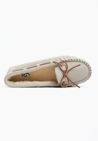 Mocasín de ante beige con un suave forro de felpa, que presenta un acento de lazo de cuero marrón y un bordado decorativo a lo largo de los bordes.