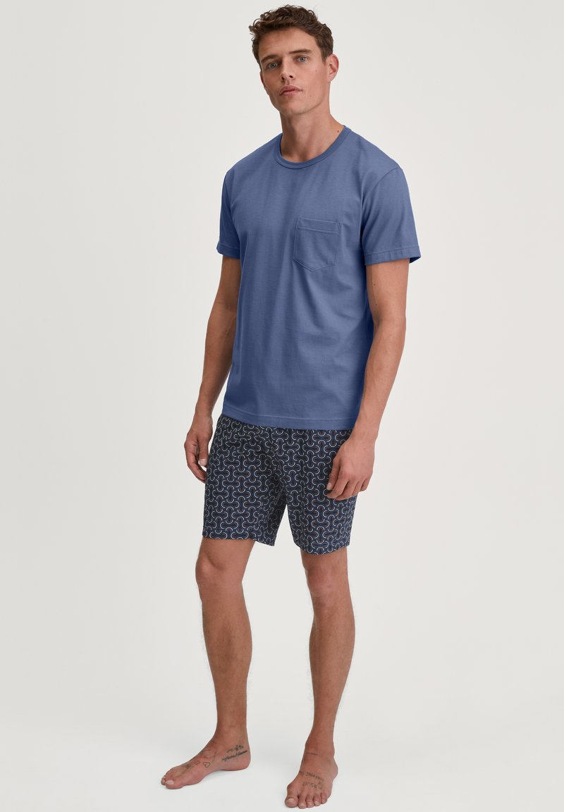 Blauwe katoenen t-shirt met een borstzak, gecombineerd met gemusterde marineblauwe shorts met witte en rode golfontwerpen. Model staat op blote voeten.