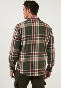 Camisa de franela a cuadros que presenta tonos verdes, rojos y beige, con mangas largas y un collar clásico con detalles de botones.