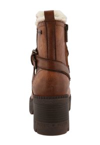 Botte cheville en cuir marron avec doublure en peluche, fermeture éclair sur le côté et sangle décorative. Présente un talon texturé et la marque "MUSTANG" sur la semelle.
