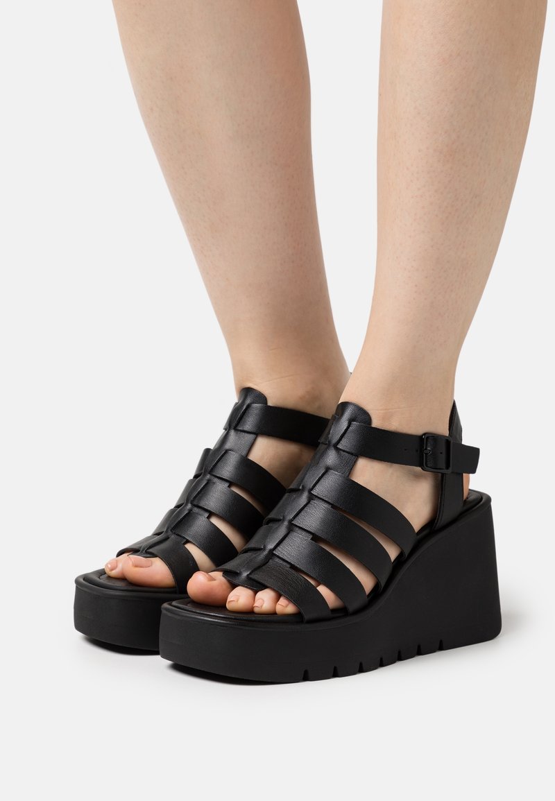 Sandales à plateforme noires en tressage avec un design compensé, une bride de cheville ajustable, une semelle texturée et un style ouvert à l'avant.