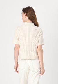 Sweter dzianinowy w odcieniu off-white z teksturowanym wzorem w diamenty, krótkimi rękawami i ściągaczem przy dekolcie, zestawiony z jasnymi spodniami.