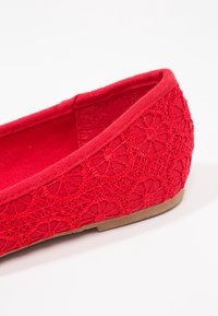 Scarpa piatta rossa con intricati motivi di pizzo circolari e suola marrone chiaro, fotografata su uno sfondo bianco.