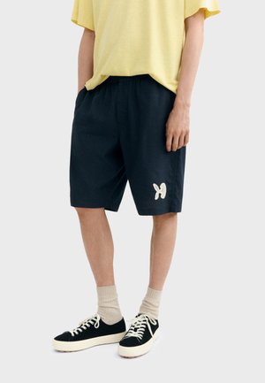 Osoba u crnim kratkim hlačama do koljena s bijelim logotipom leptira, žutom majicom, bež čarapama i crnim tenisicama s bijelim vezicama.