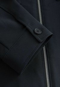 Schwarze Stoffjacke mit silbernem Reißverschluss, ausgestattet mit einem geknöpften Bündchen und glatter Textur. Das Design ist tailliert mit klaren Linien.