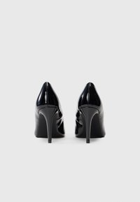 Un paio di scarpe con tacco alto in vernice nera lucida viste da dietro su uno sfondo bianco uniforme.