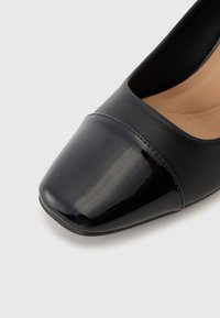 Scarpa con tacco alto in pelle verniciata nera, con punta quadrata, caratterizzata da una finitura liscia e dettagli opachi. Design minimalista con una soletta neutra.