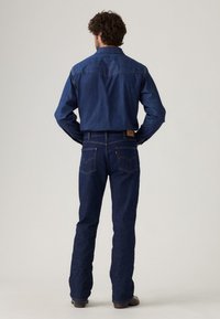Camisa de mezclilla oscura y jeans llevados por una persona, con un corte clásico, detalles de costura y un parche de cuero de la marca en la parte trasera.