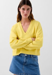 Felpa gialla con scollatura a V, polsini e orlo a costine; abbinata a una mini gonna di jeans. Tessuto morbido e vestibilità informale.
