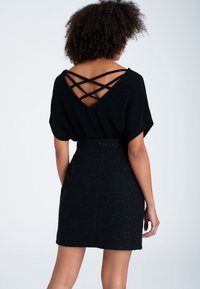 Žena s kudrnatými vlasy, oblečená v černém topu s křížovými popruhy na zádech a texturované černé sukni, stojící proti jednoduchému pozadí.