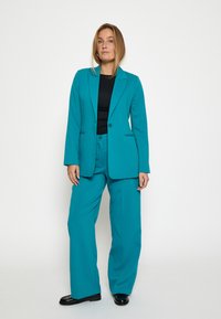 Tyrkysový oblek s přiléhavým sakem s klopami a kalhotami širokého střihu. Materiál vypadá hladce s mírnou strukturou. Černý top pod ním.