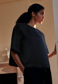 Žena s kudrnatými vlasy v culíku, oblečená v volném černém topu, stojící uvnitř u dveří, s jednou rukou v kapse.