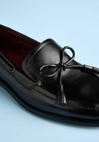 Schwarzer Leder-Slipper mit abgerundetem Zehenbereich, ausgestattet mit einer Schleifen- und Quastenverzierung. Glatte Textur und Nahtdetails an der Seite.