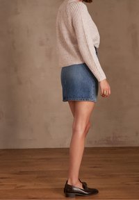 Kvinna i en beige stickad tröja, blå denim mini kjol, svarta loafers och en stor ring, står på ett trägolv mot en beige vägg.