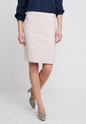 Kvinde iført en lysebeige nederdel til knæet, marineblå prikket bluse og højhælede sko med slangeprint, stående med krydsede ben.