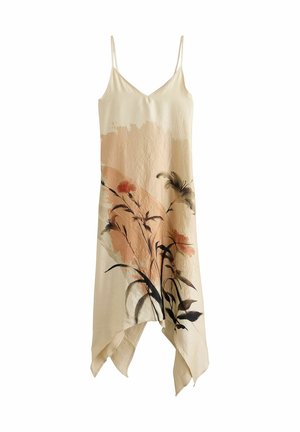 Kvinde med langt mørkt hår iført en ærmeløs beige kjole med blomsterprint, stående på stentrapper mod en struktureret stenvæg.