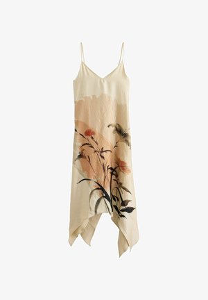 Frau mit langen dunklen Haaren, die ein ärmelloses beiges Kleid mit Blumenmuster trägt, steht auf Steinstufen vor einer strukturierten Steinmauer.