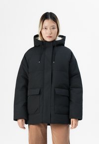 Kvinne som har på seg en svart hettejakke med pufferdesign og frontlommer, samt beige kordfløyelsbukser, stående mot en hvit bakgrunn.