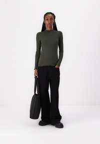 Long-sleeve, ribbed top in olive green, paired with wide black pants and a black tote. Model stands on a plain white background.