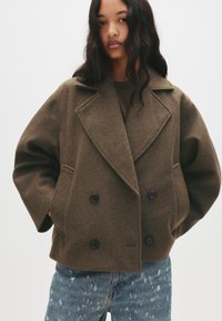 Brown wool blend double-breasted coat with large lapels and two front pockets, paired with distressed blue jeans.