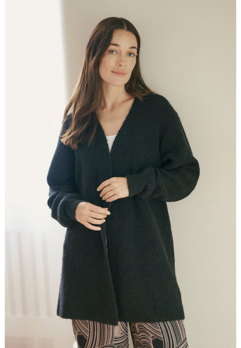 Zwarte gebreide cardigan met een V-hals, lange mouwen en een open voorkant. Heeft een gestructureerde afwerking en valt tot halverwege de dij.