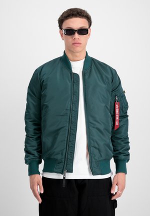 Zöld bomberdzseki sima, fényes textúrával, cipzárral, bordás mandzsettákkal és piros címkével a ruhán. Fehér pólóval viselve.