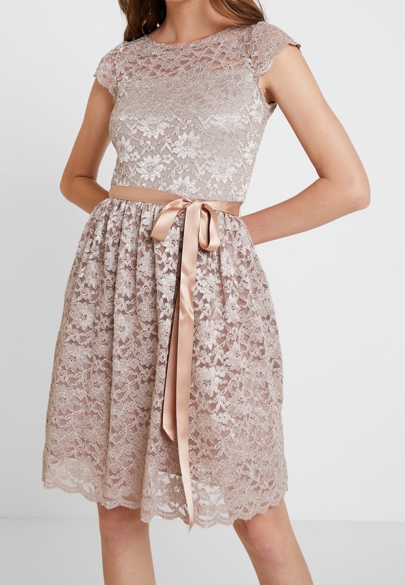 Robe sans manches en dentelle rose pâle avec motif floral, corsage ajusté, ourlet festonné, et ruban en satin noué en un nœud à la taille.