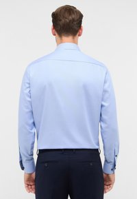 Camicia a maniche lunghe azzurro chiaro con colletto classico, texture liscia e polsini con bottoni, indossata con pantaloni scuri. Design semplice e su misura.