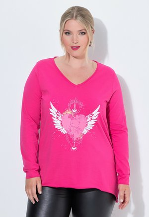 Femme aux cheveux blonds portant un haut rose vif à manches longues avec un motif de cœur ailé et un pantalon noir, debout devant un fond blanc.