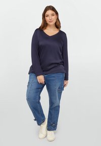 Marineblauwe longsleeve top met V-hals, gecombineerd met blauwe cargobroek met meerdere zakken en witte sneakers. Glad materiaal en een relaxte pasvorm.