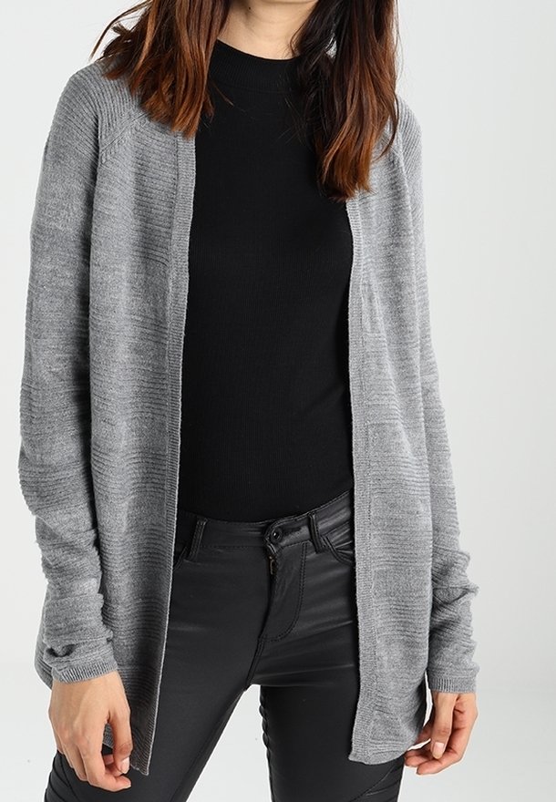Femme portant un cardigan gris à manches longues sur un haut noir et un pantalon noir, se tenant devant un fond blanc uni.