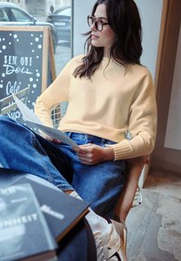 Mujer joven con gafas, vestida con un suéter amarillo y jeans, sentada junto a la ventana en una cafetería, leyendo el menú, con libros en la mesa cercana.