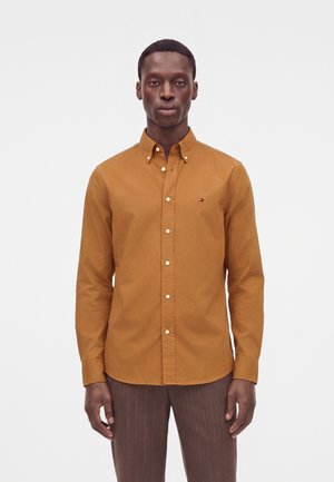Homme portant une chemise moutarde boutonnée avec un petit logo sur la poitrine et un pantalon marron à fines rayures, debout devant un fond blanc.