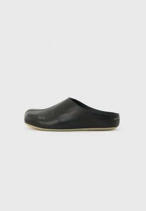 Zapato de cuero negro tipo slip-on con punta cerrada y parte trasera abierta, suela beige baja y costuras mínimas sobre un fondo claro y liso.