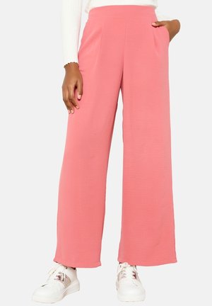 Femme portant un pantalon rose taille haute à jambes larges avec des poches sur les côtés et des baskets blanches, debout avec une main dans la poche.