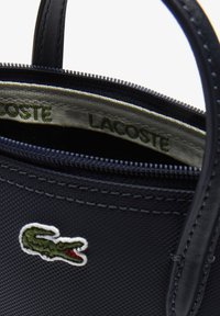 Sac à main bleu marine en matériau texturé, fermeture éclair, et une marque Lacoste avec un logo crocodile vert brodé.