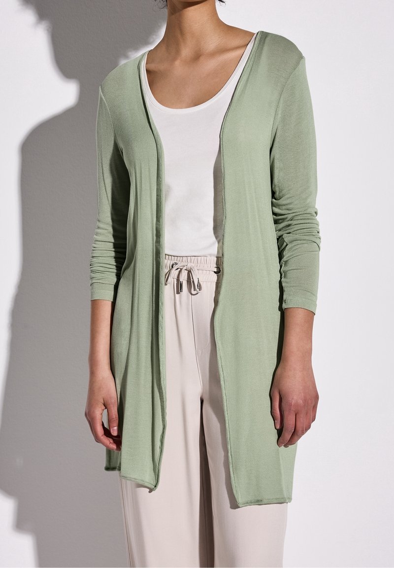 Personne portant un cardigan vert clair ouvert sur un haut blanc à col rond et un pantalon beige à cordon, debout devant un fond uni.
