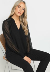 Blusa negra con escote en V y diseño de lunares texturizados. Mangas largas transparentes con puños fruncidos. Combinada con pantalones negros, sentada en una silla.